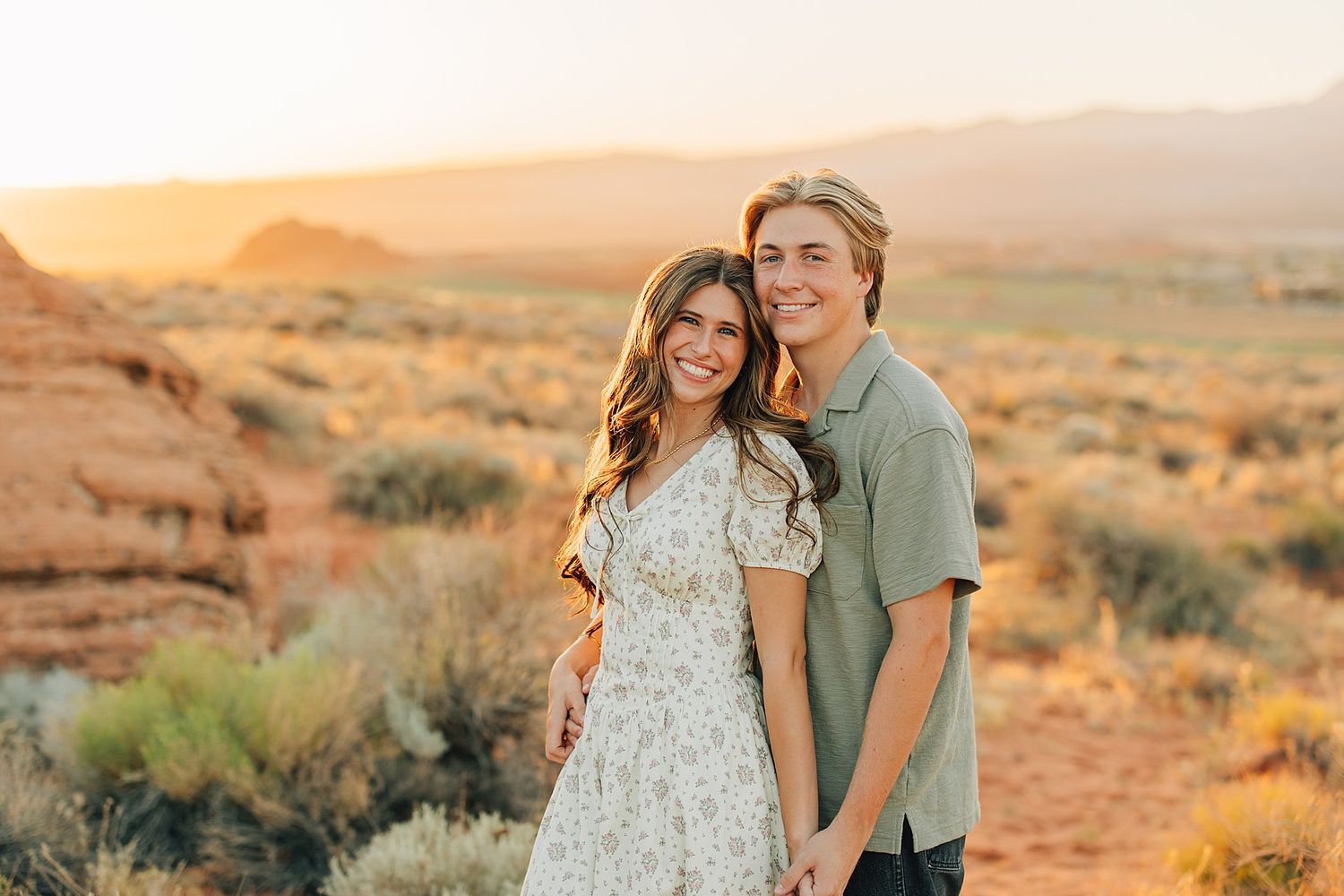 Couple smiling at sunset in a desert landscape with red rocks and mountains in the background.