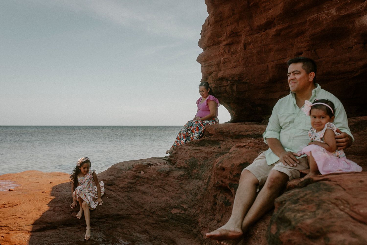 Family sitting on red sandstone rocks by the ocean shore, with calm sea and cloudy sky in the background.