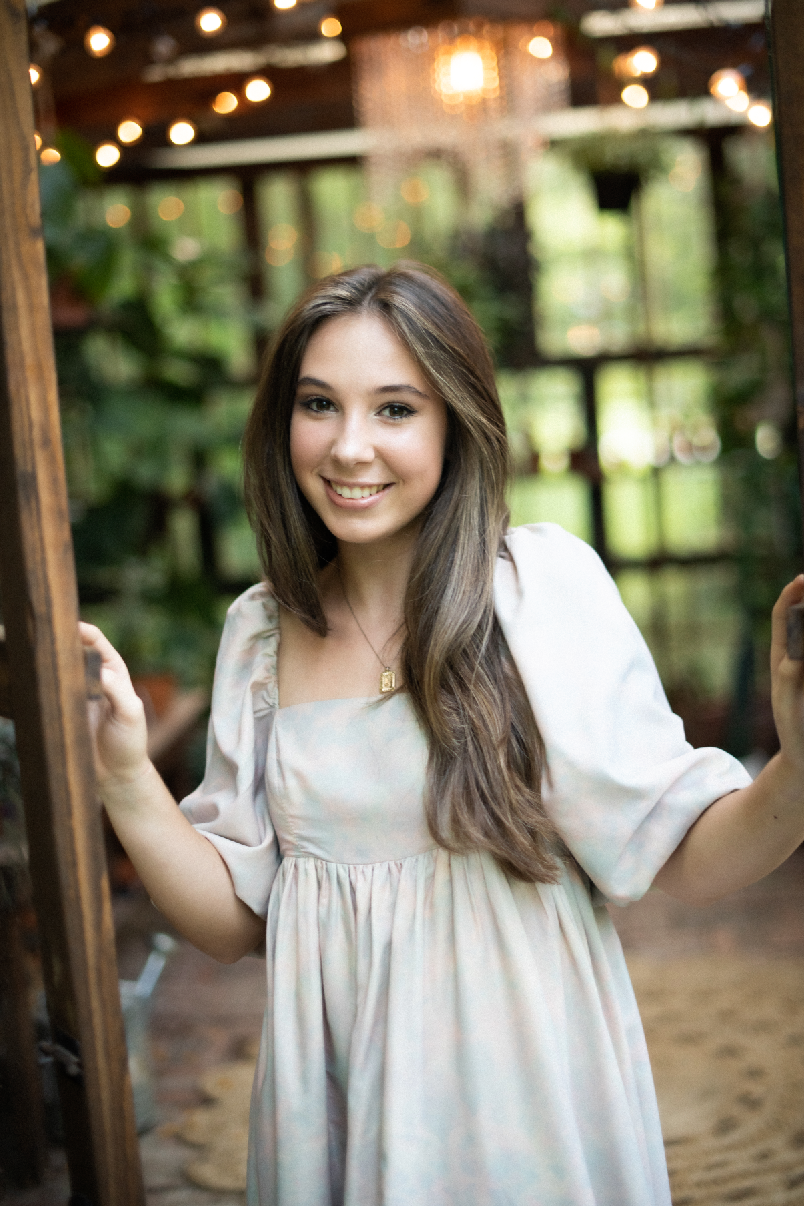 Young Black woman in white top smiles outdoors, wearing gold jewelry, with green willow tree background.