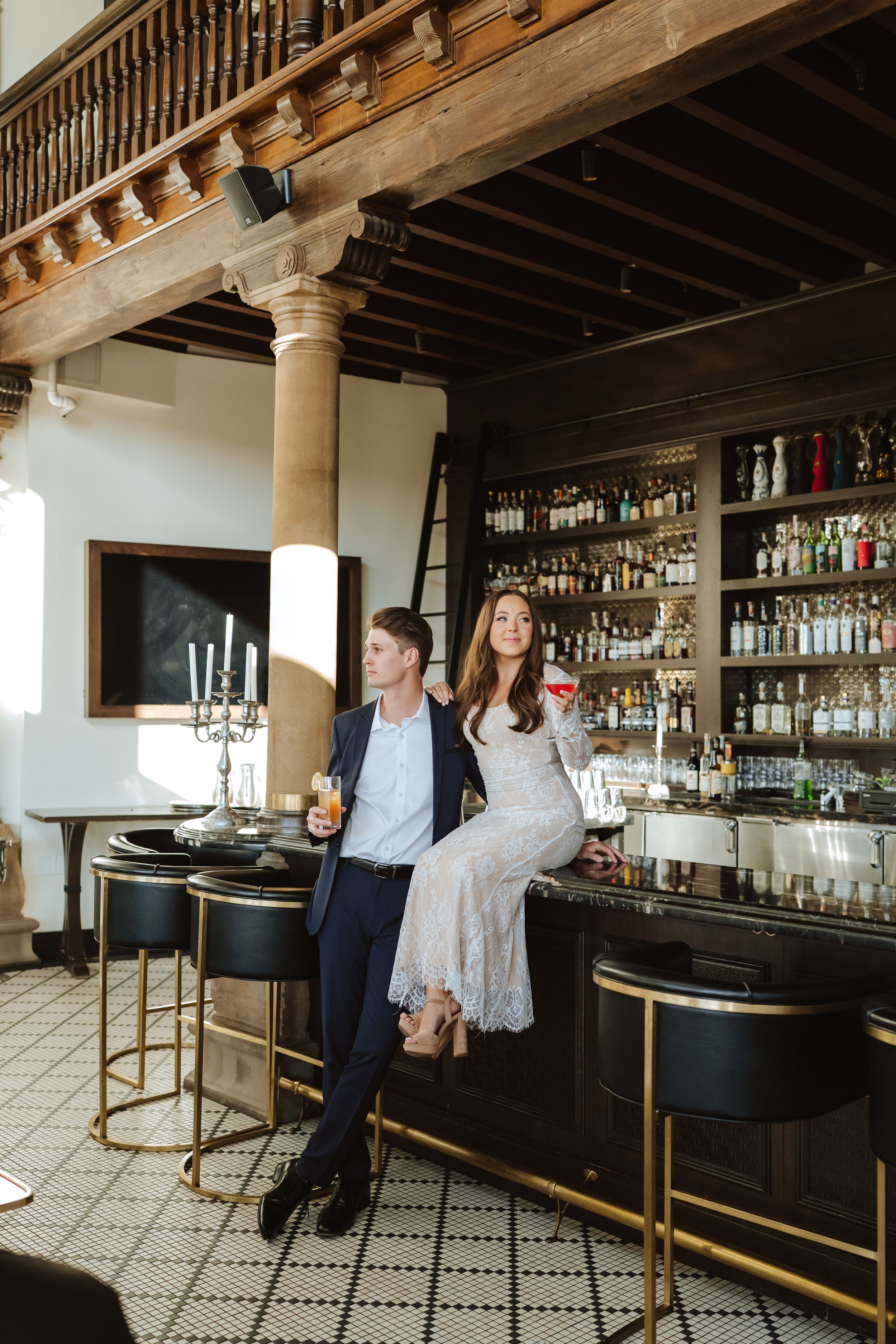 A couple sits on the elegant bar holding cocktails at Wrigley Mansion. Groom wears a blue suit jacket and bride wears a lace fitted dress