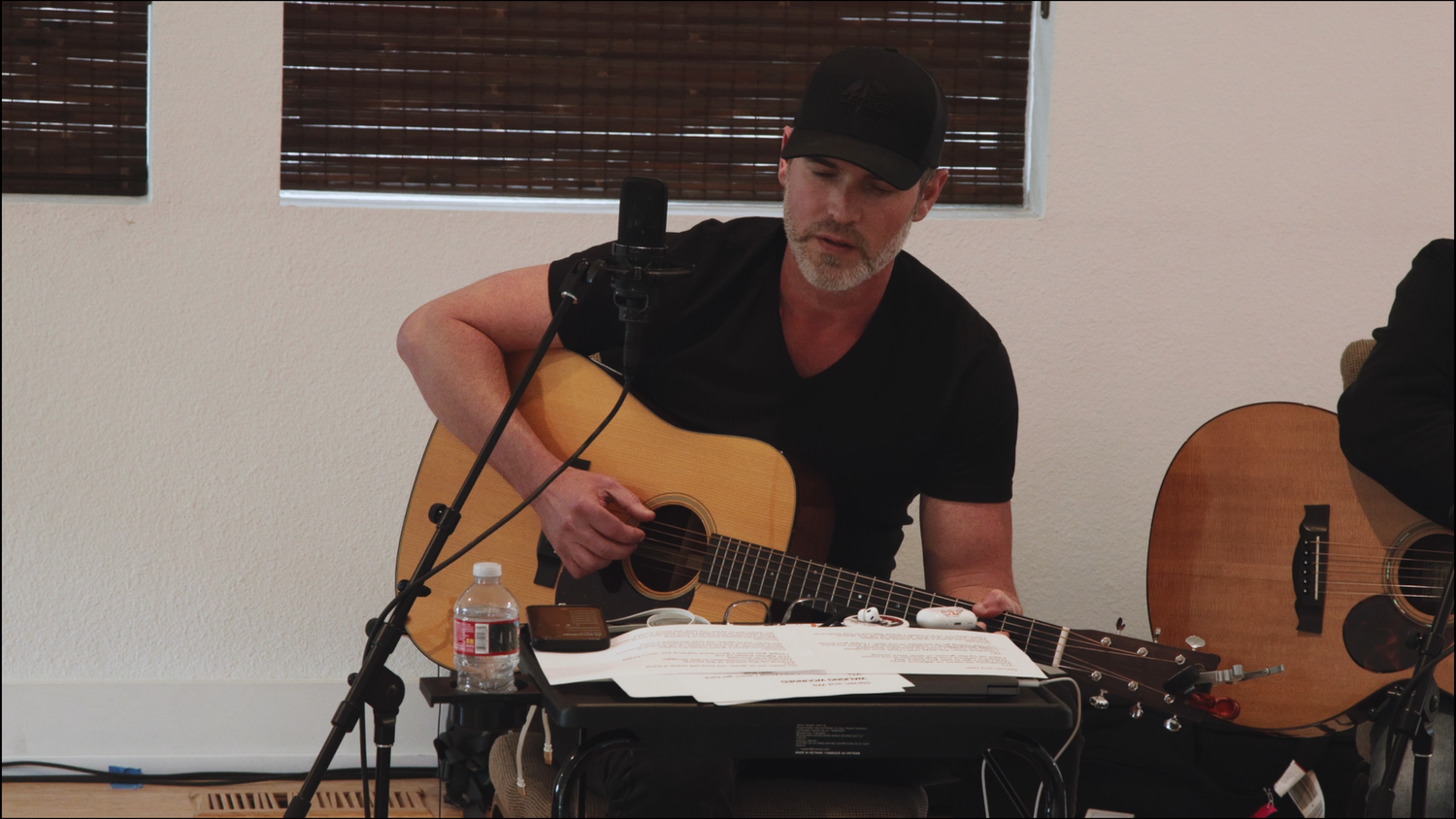 Man in black cap playing acoustic guitar at microphone stand with sheet music and water bottle nearby.