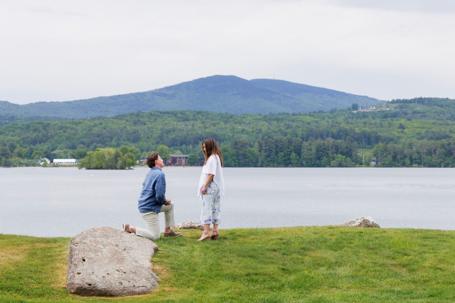 Man proposing to woman by a serene lake with mountain views on a lush green lawn.