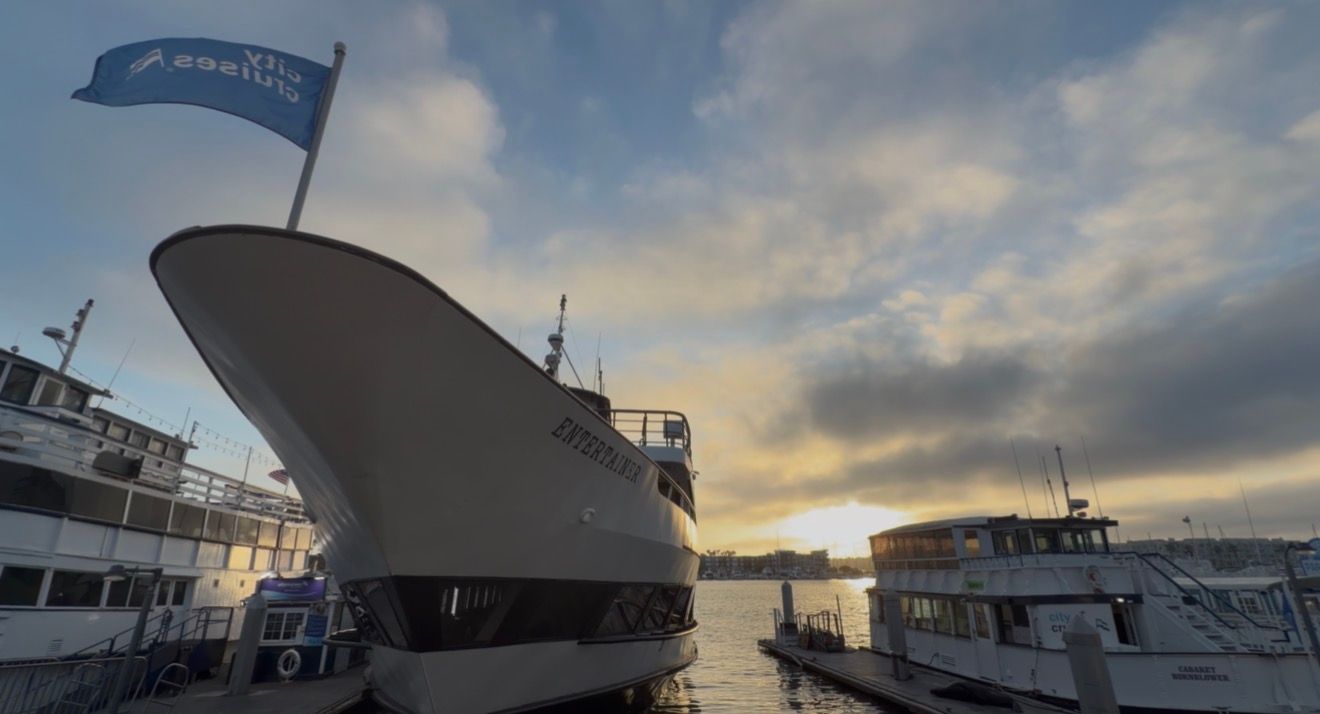 Large white cruise ship docked at marina during sunset with dramatic cloudy sky reflected on calm water.