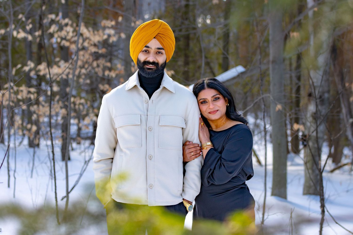 A couple stands together in a snowy winter forest setting wearing coordinated white and dark outfits.