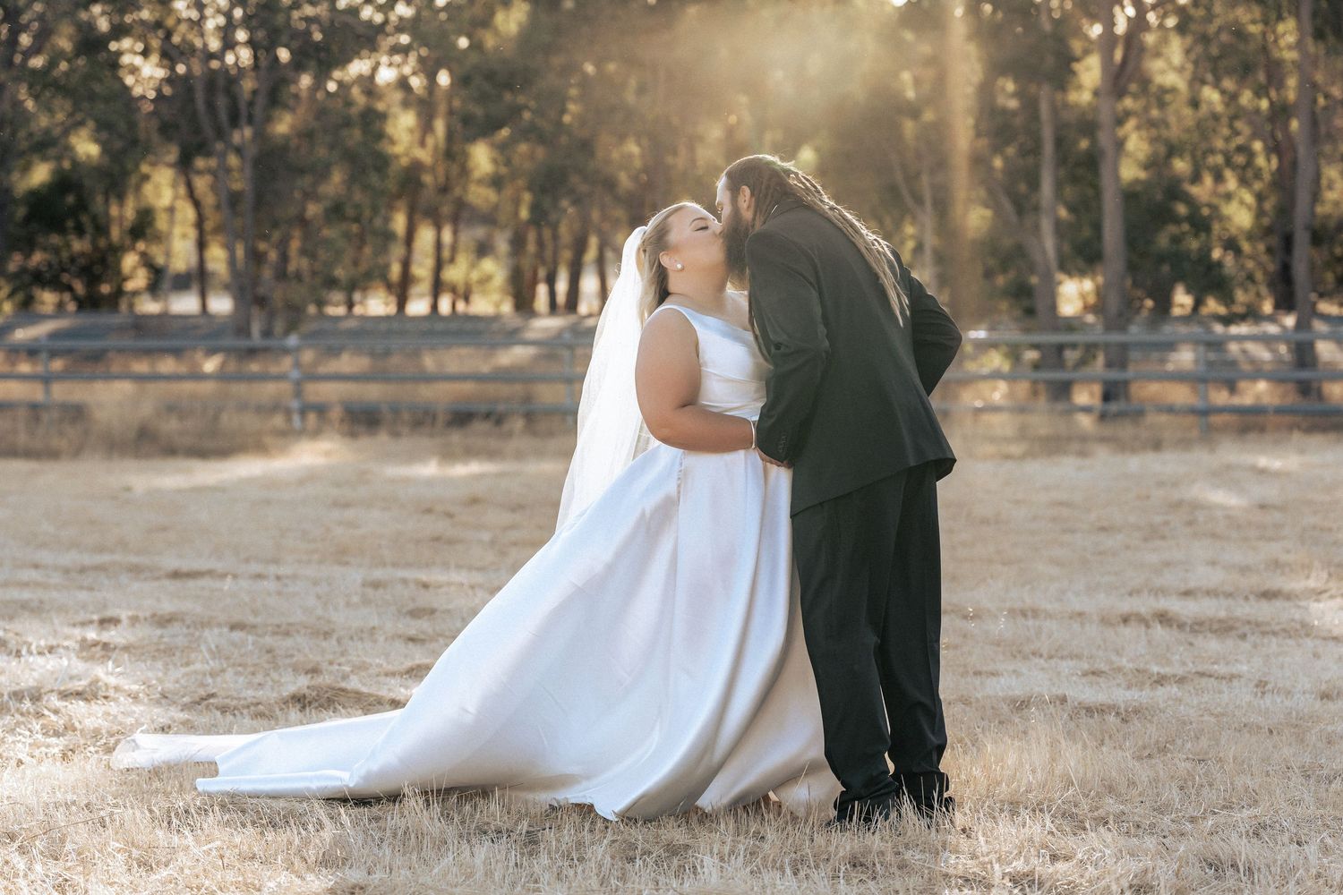 Bride and groom sharing a romantic kiss in a sunlit field with trees and a fence in the background.