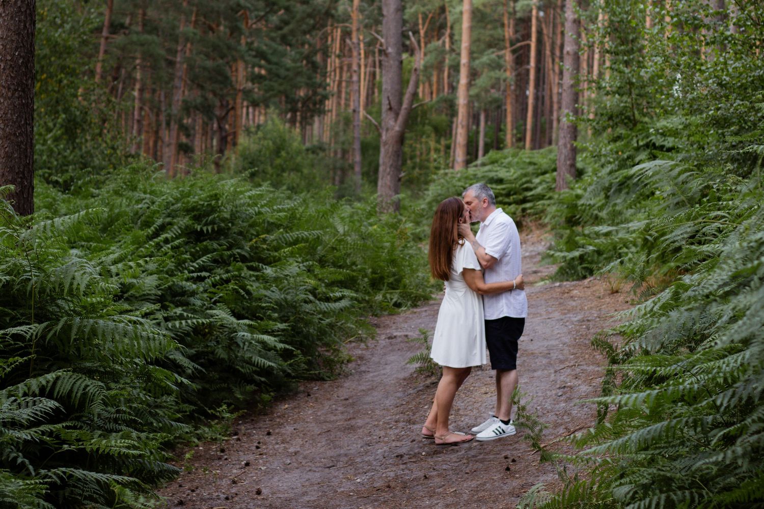 A couple embraces on a forest path surrounded by lush ferns and tall pine trees in a romantic outdoor setting.