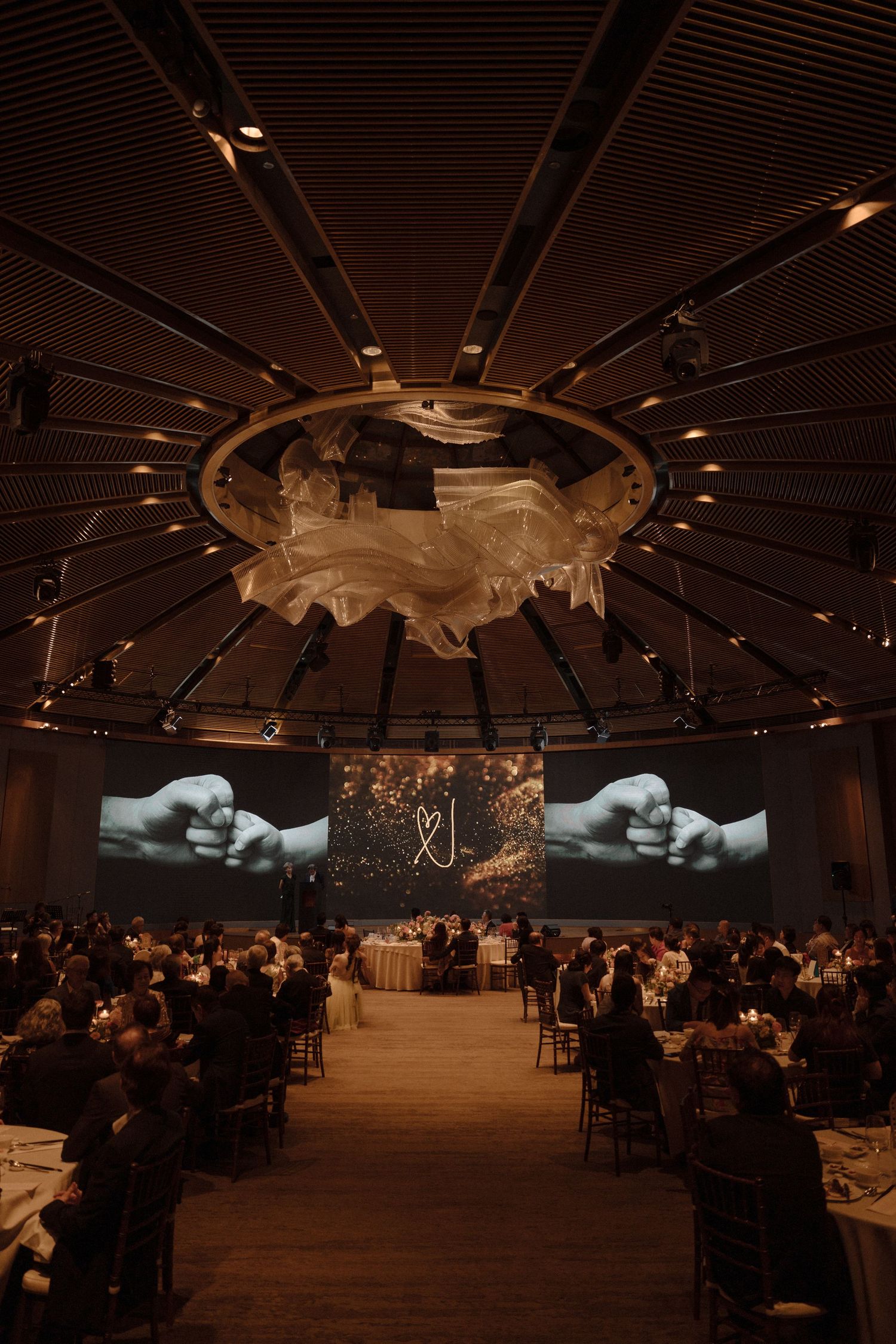 Elegant wedding reception in grand ballroom with large screens displaying couple's monogram and guests seated at candlelit tables.