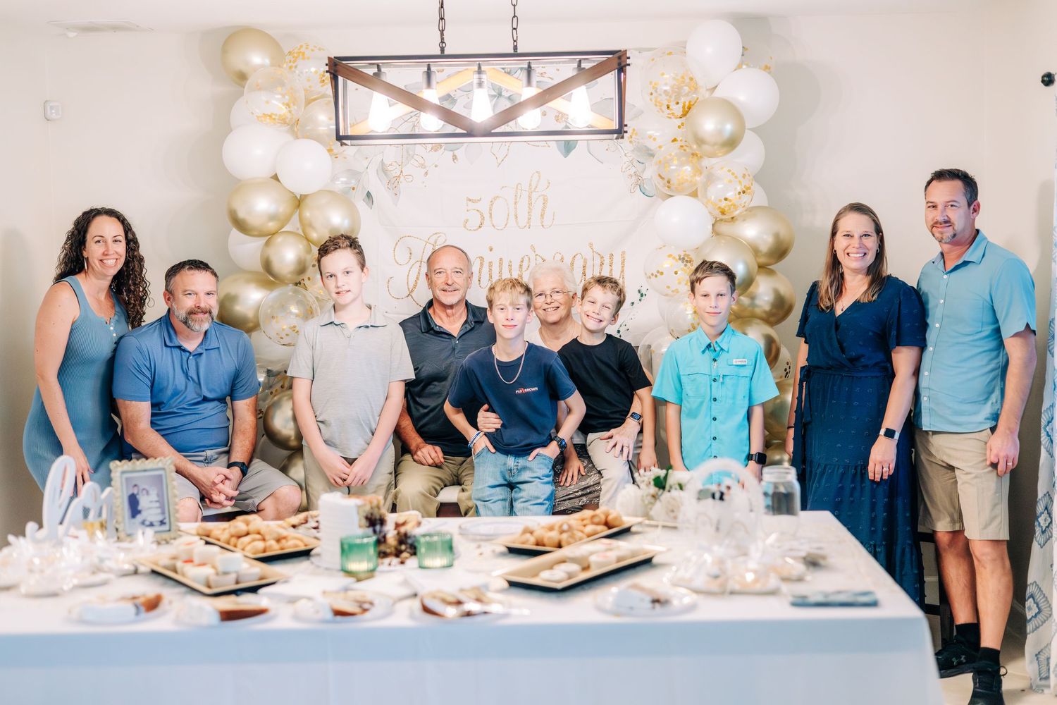 Family gathering around a decorated table at a 50th anniversary party with gold and white balloon arch backdrop.