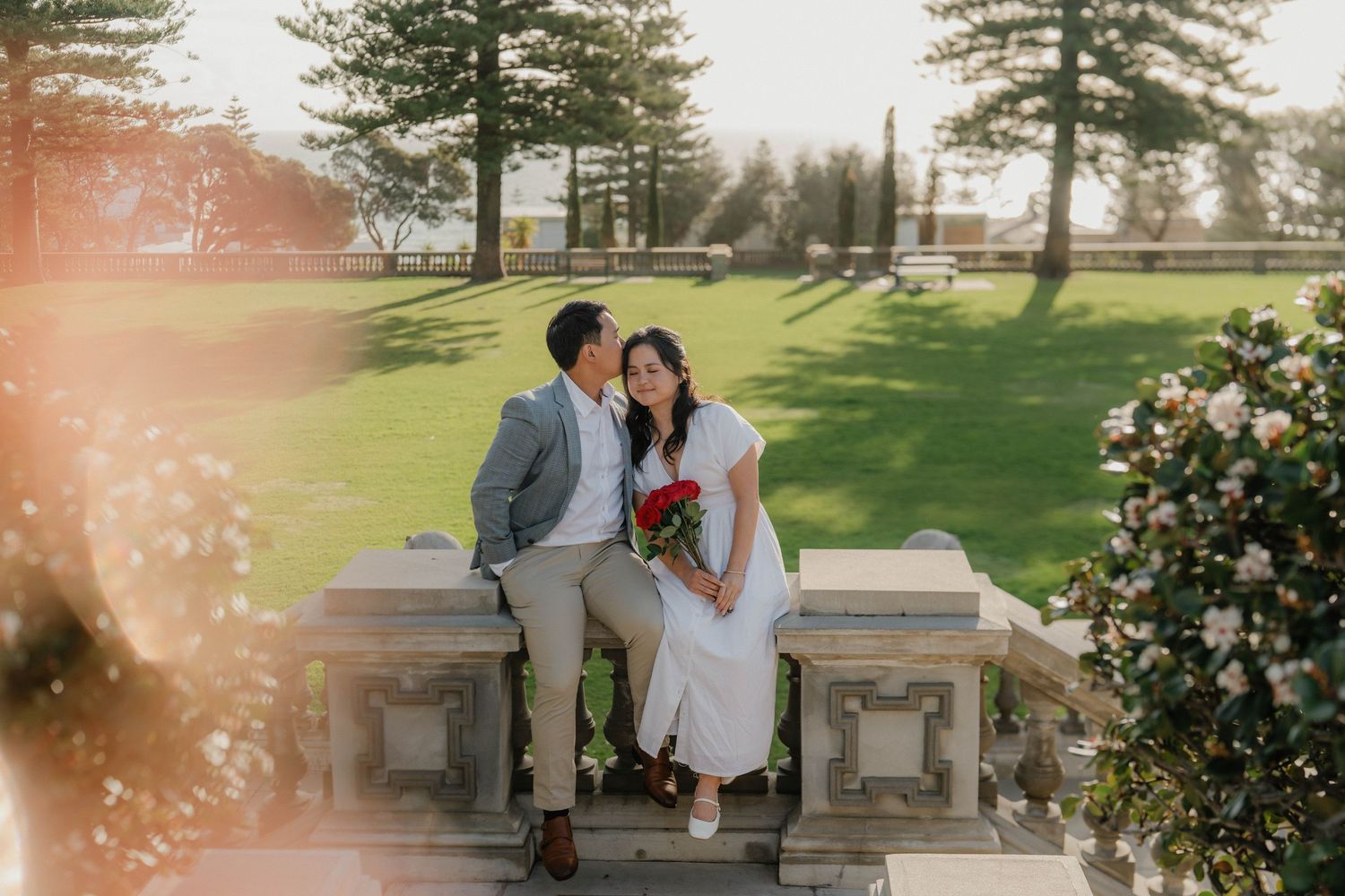 A couple shares an intimate moment on a stone bench in a sunlit garden with blooming flowers and evergreen trees.