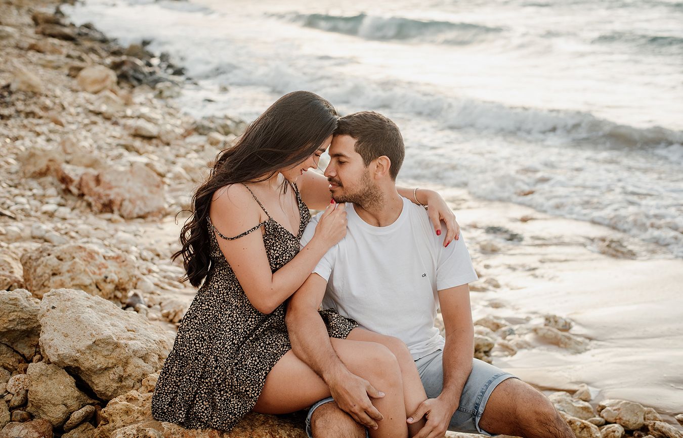 Romantic couple embracing on a sandy beach with ocean waves, rocky cliffs in the background at golden hour.