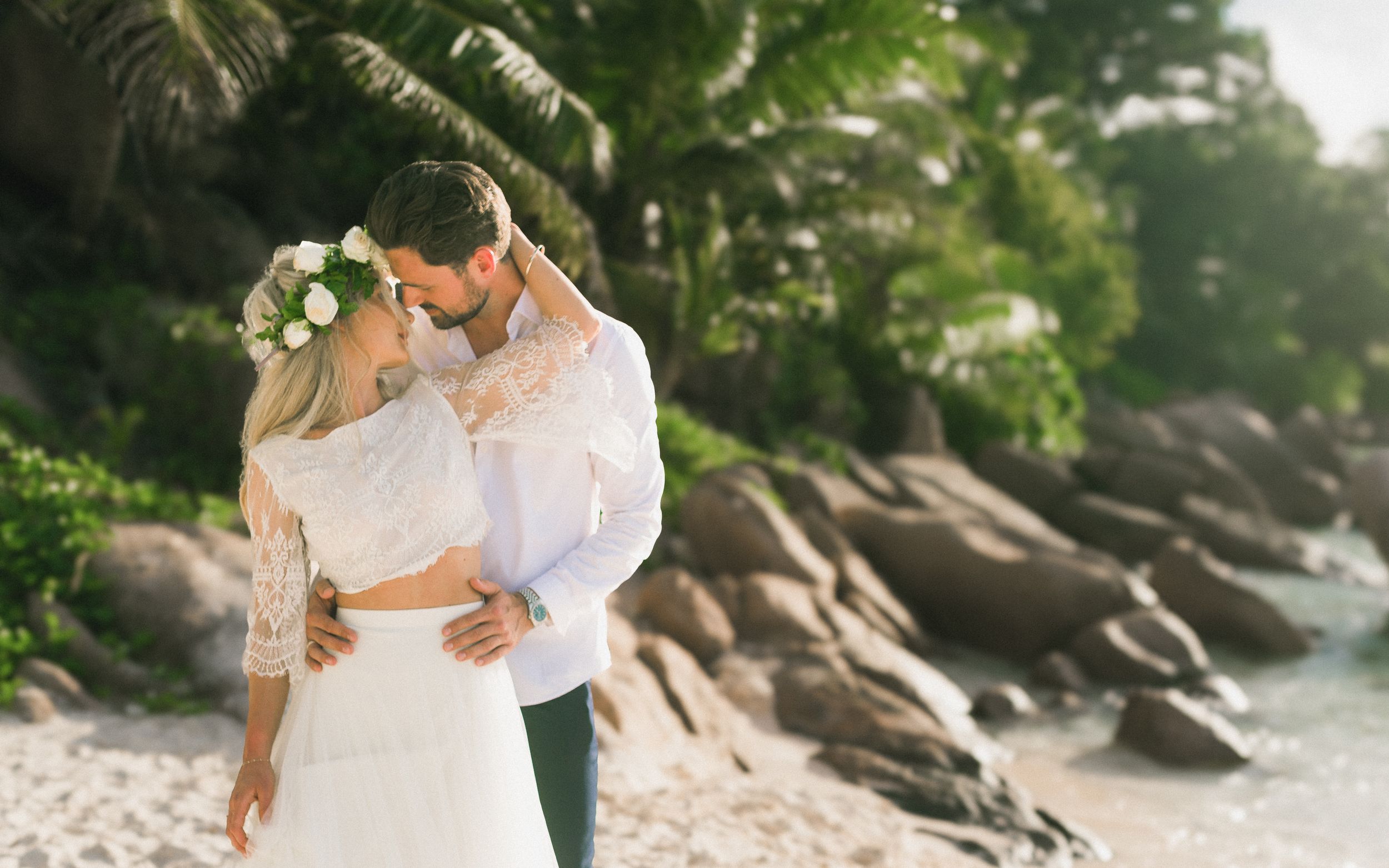 Couple in wedding attire sharing a romantic kiss on a tropical beach with lush greenery and rocks.