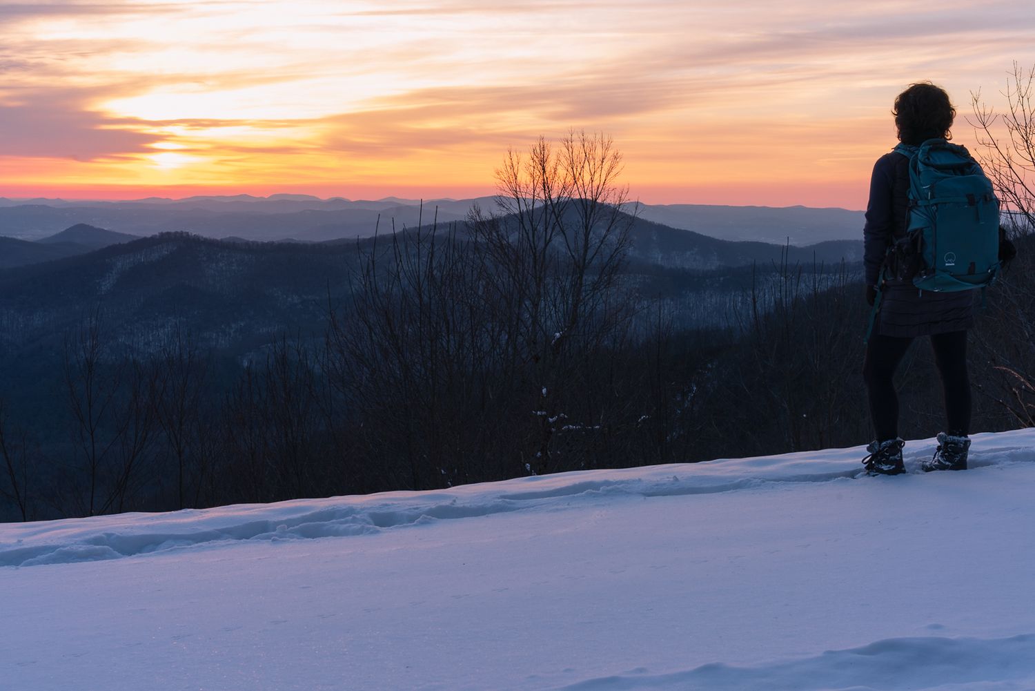 Hiker with backpack stands on snowy mountain overlook watching a colorful winter sunset over rolling forested hills.