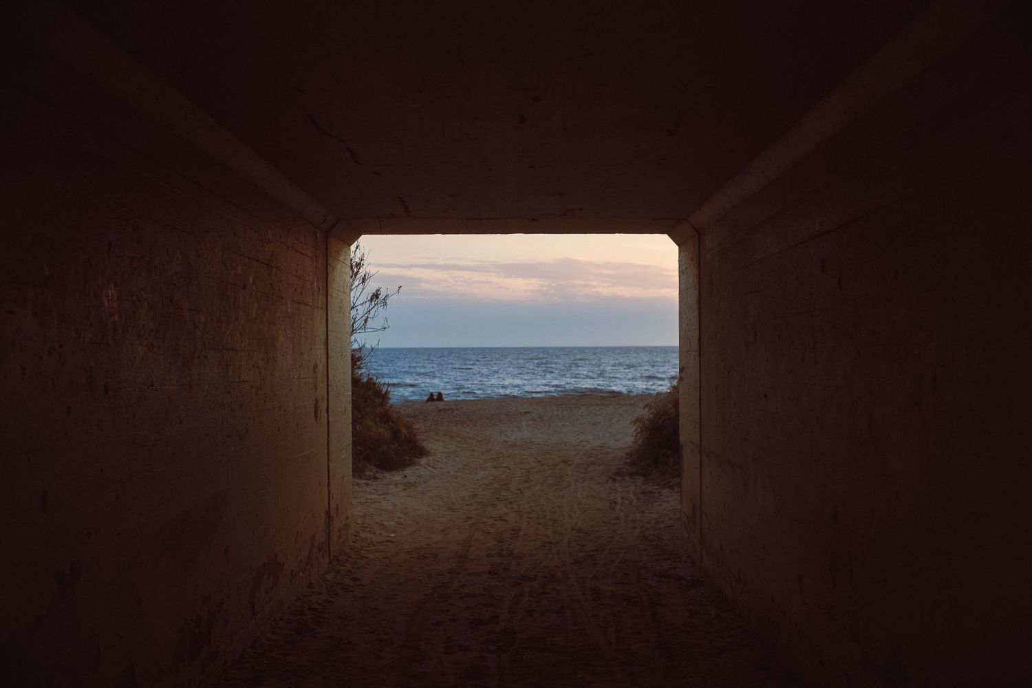 View through a dark concrete tunnel opening onto a sandy beach with calm ocean waves at dusk.