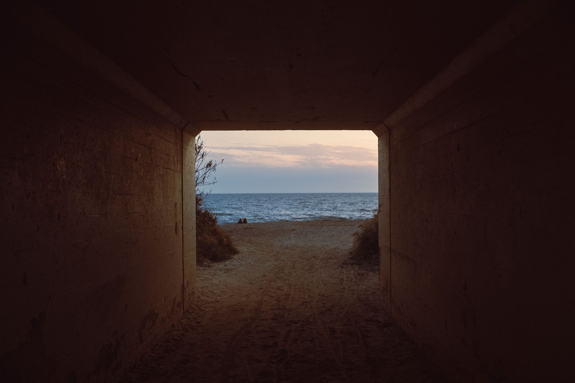 View through a dark concrete tunnel opening onto a sandy beach with calm ocean waves at dusk.
