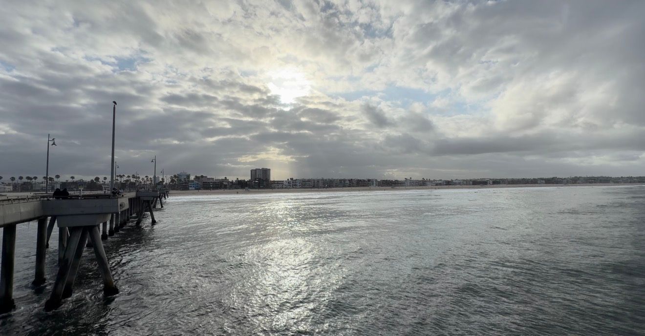 Scenic pier view over calm ocean waters with dramatic cloudy sky and city skyline at sunset.