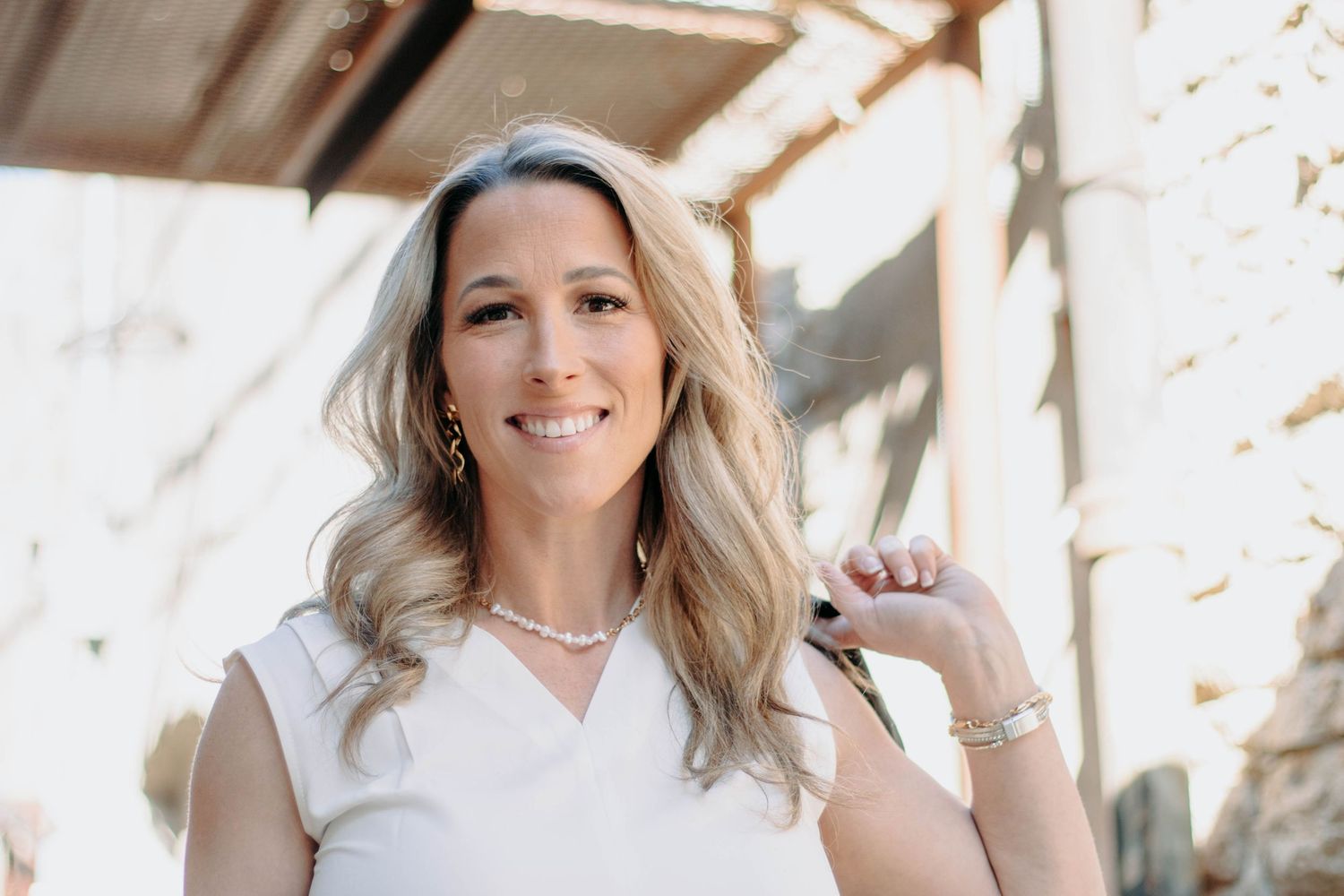 Smiling blonde woman in white top wearing pearl necklace and gold earrings, posing outdoors in natural light.