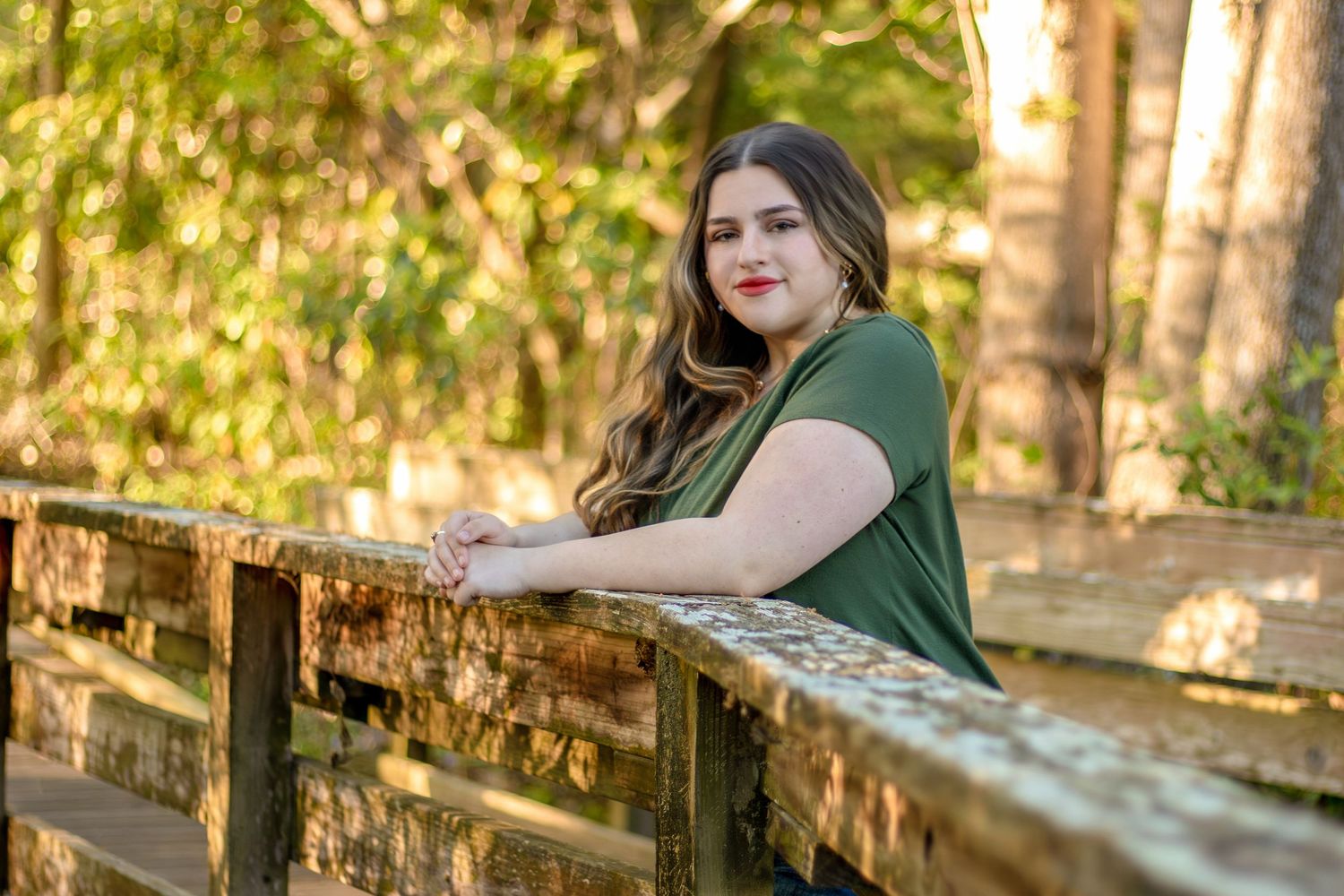 Young woman leaning on wooden bridge railing surrounded by trees in golden light. family photography, graduate sessions, portrait weddings