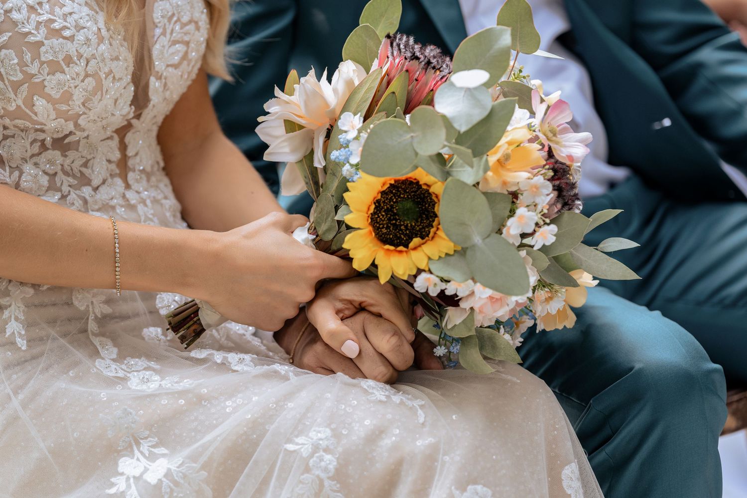 Bride and groom holding a wedding bouquet featuring a sunflower, eucalyptus, and pastel blooms.