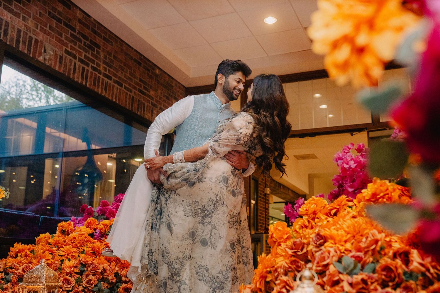 Couple sharing a romantic embrace surrounded by vibrant orange and pink floral arrangements at an Indian wedding celebration.