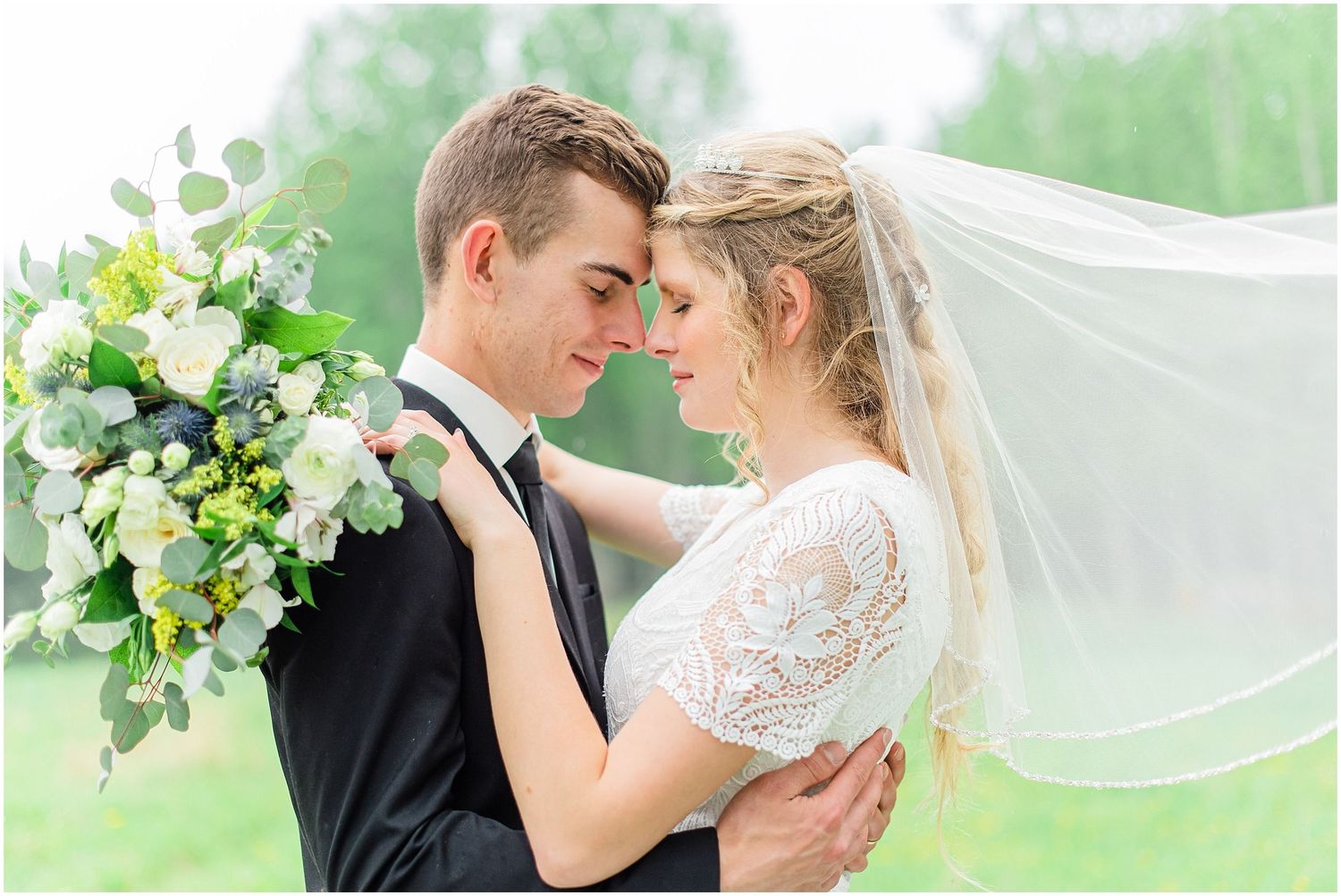 Bride and groom touching foreheads romantically outdoors, her veil flowing in the breeze, holding a lush green and white bouquet.