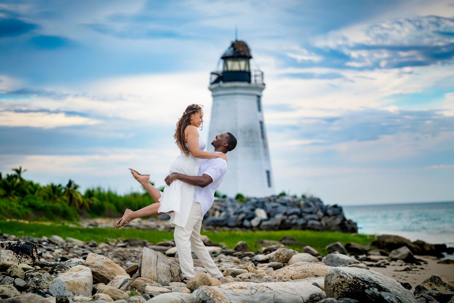 Couple embracing by a white lighthouse on rocky shore with ocean and cloudy sky backdrop.