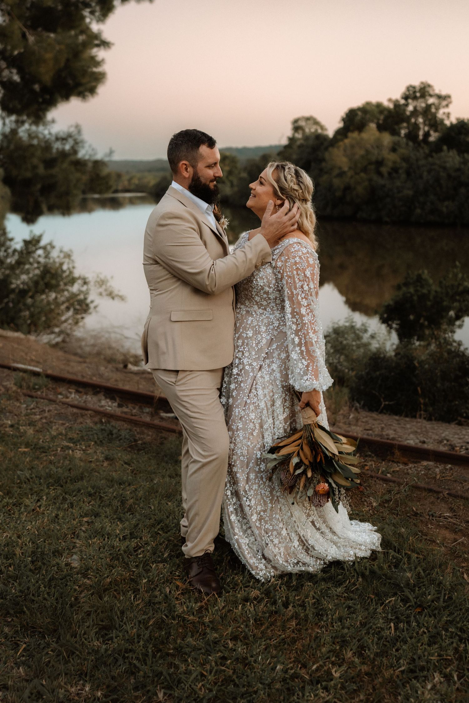 Couple sharing a romantic moment at sunset by a river, groom in tan suit, bride in sequined gown holding a bouquet.