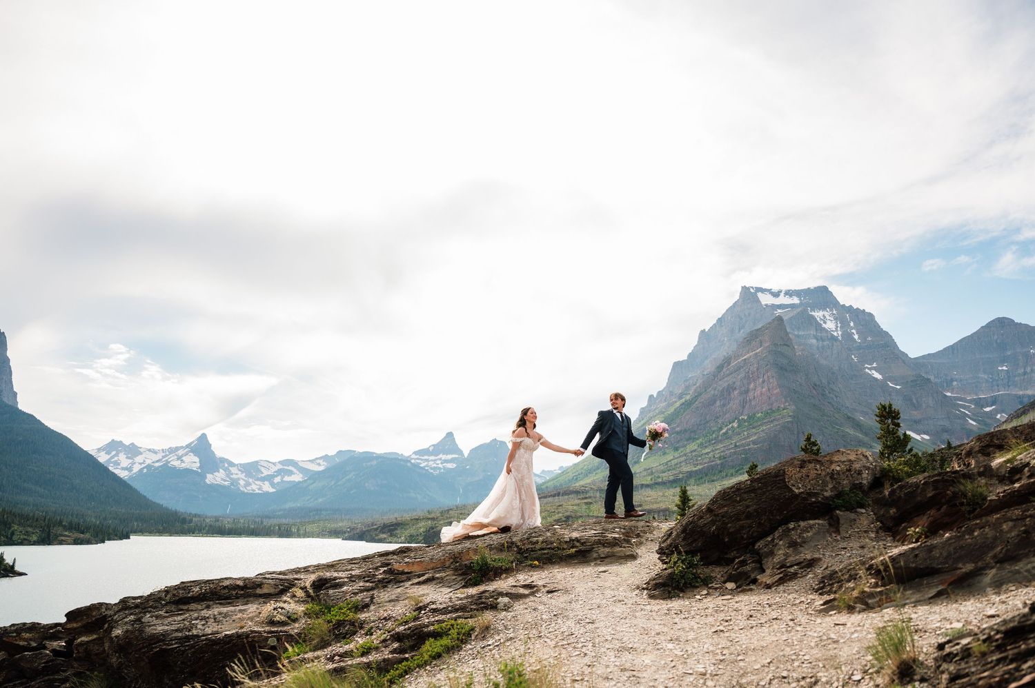 Bride and groom hold hands on rocky mountain overlook with stunning lake and glacier peaks backdrop.