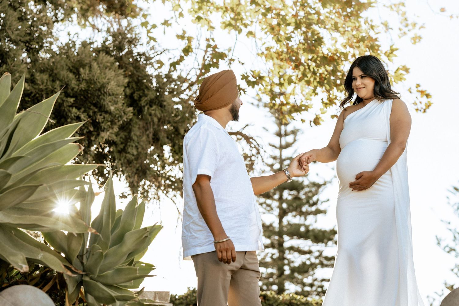 A pregnant woman in a white dress holds hands with a man outdoors during a maternity photoshoot.