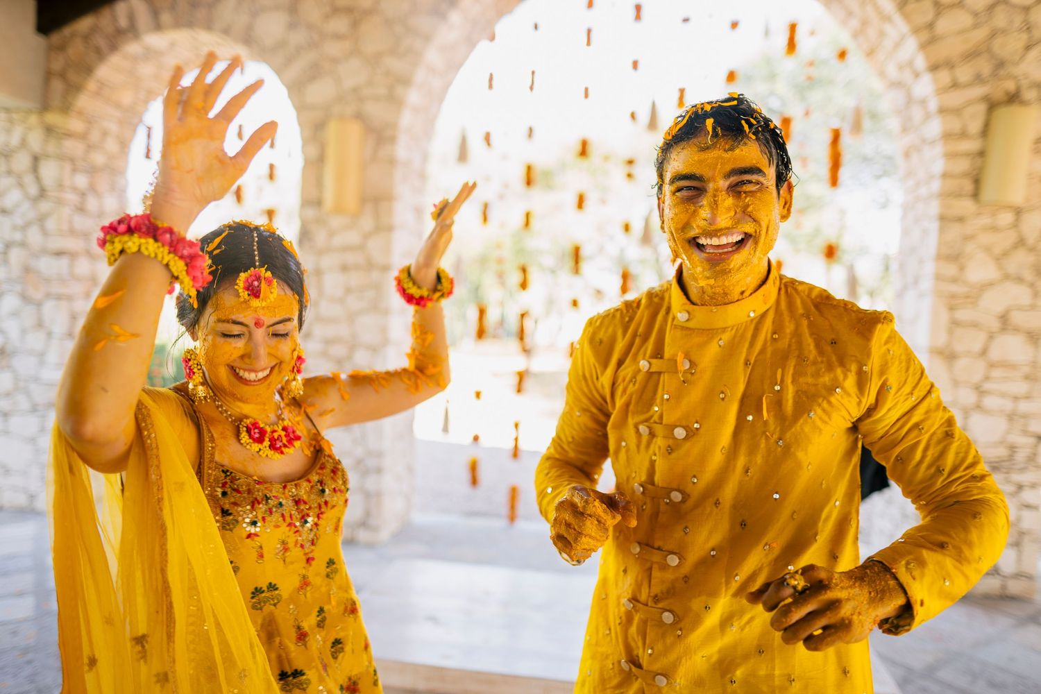 Joyful Indian couple in matching yellow outfits celebrating Haldi ceremony with colorful decorations.