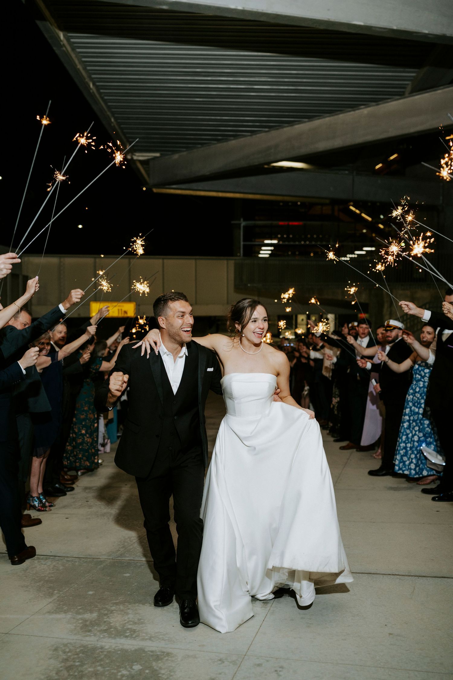 Bride and groom walk through sparkler exit surrounded by cheering wedding guests at night.