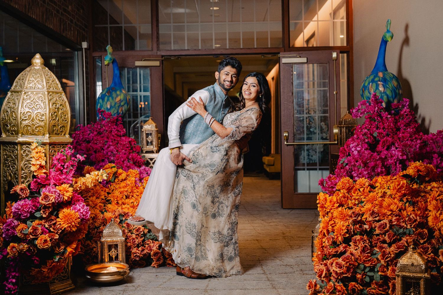 A person poses in a gray gown against a backdrop of vibrant pink and orange flowers in an elegant indoor setting.