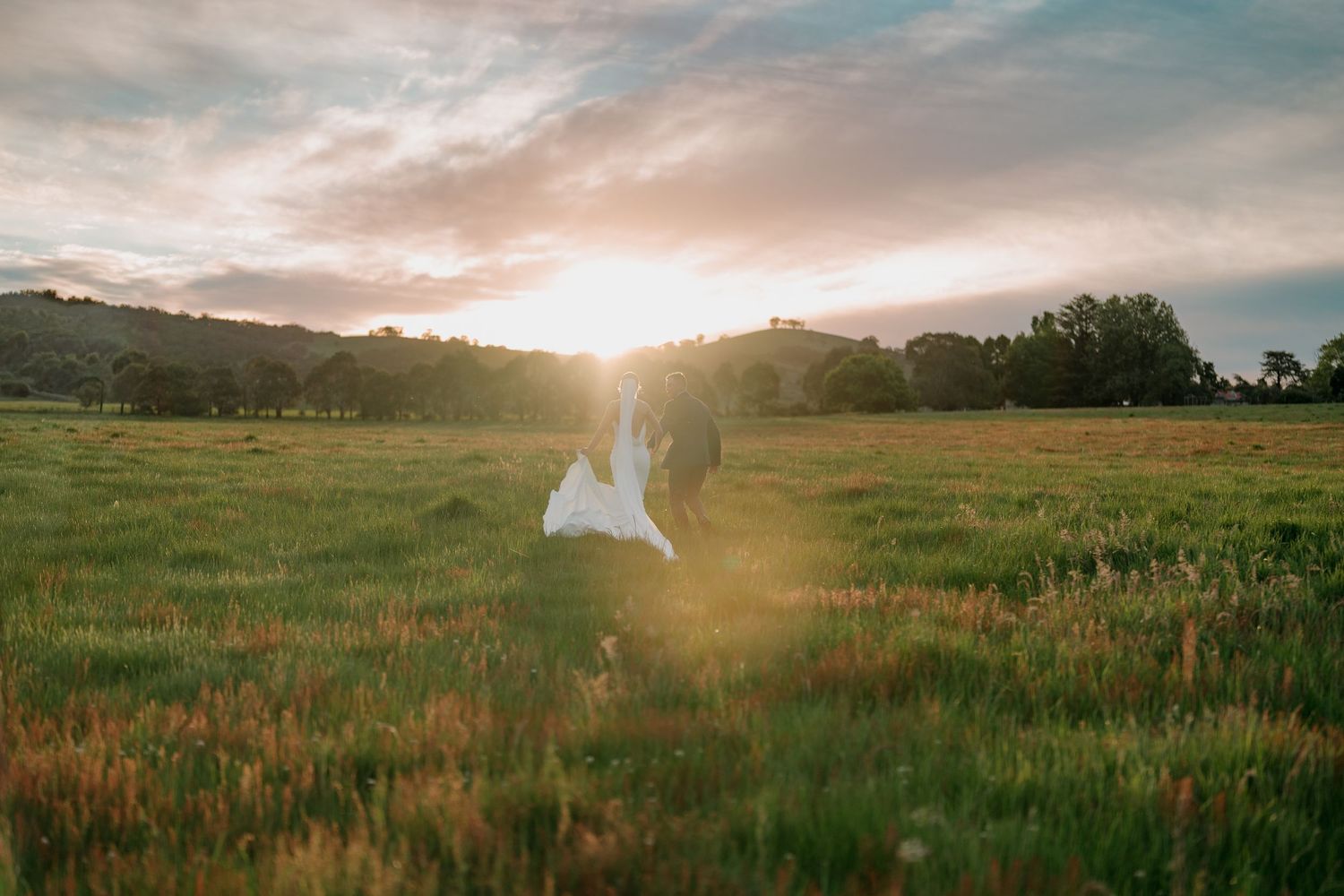 Bride and groom walk through a sunlit green field at golden hour with rolling hills and dramatic sky.