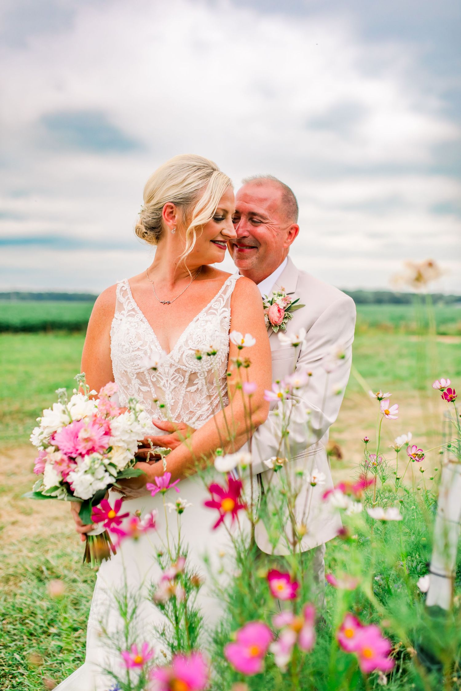 Bride and groom share a romantic moment in a wildflower field, bride holding pink and white bouquet in white lace gown.