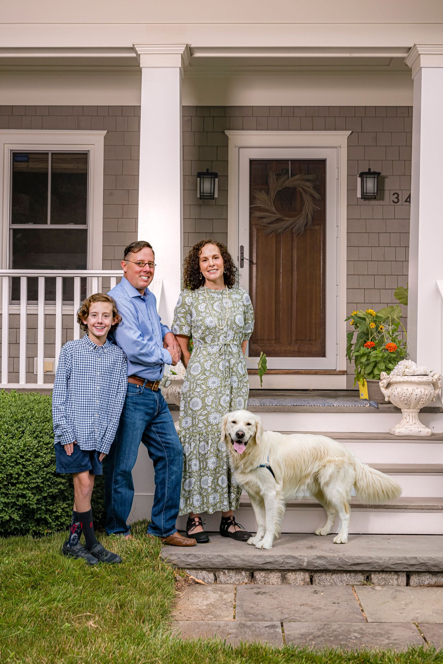 Family of three with golden retriever posing on stone steps outside gray shingle home with white porch columns.