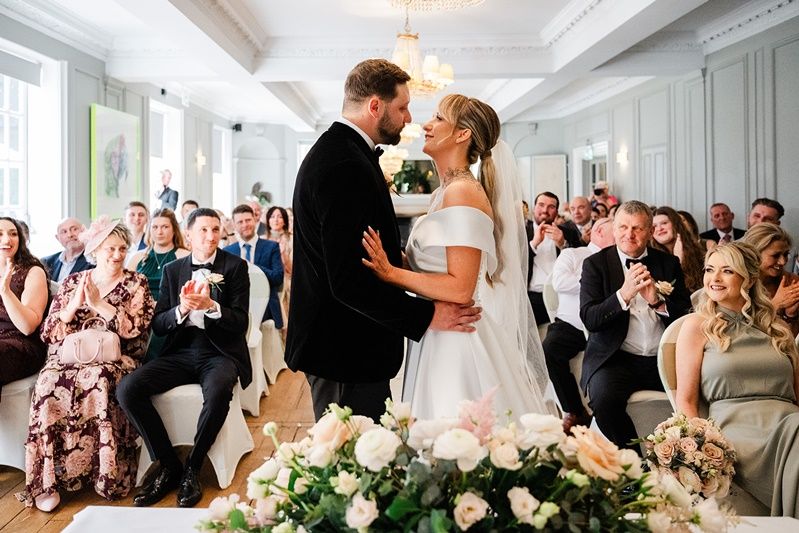 Bride and groom share first kiss at elegant wedding ceremony with guests applauding in decorated reception hall.