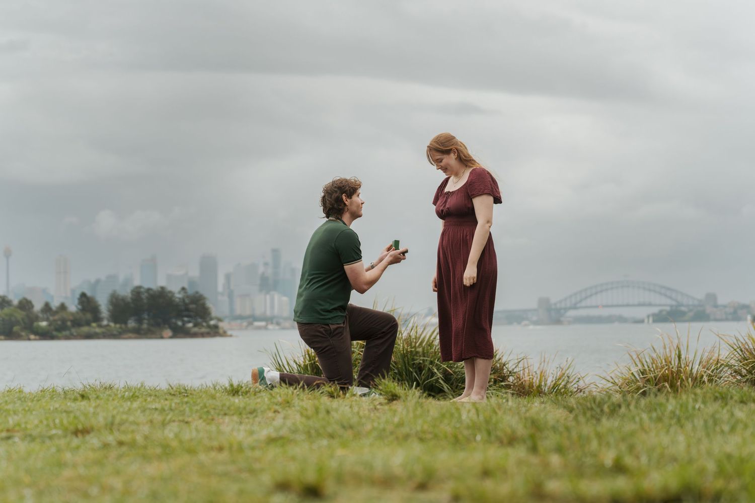Man proposing to woman on grassy waterfront with Sydney Harbour Bridge and city skyline in background.