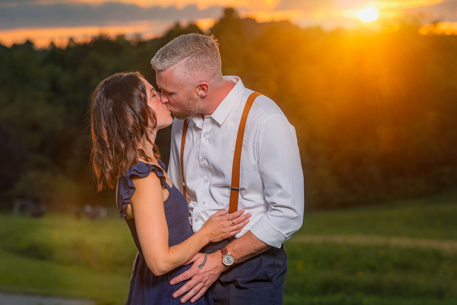A couple shares a romantic kiss during a golden sunset in a lush outdoor setting with warm lighting.
