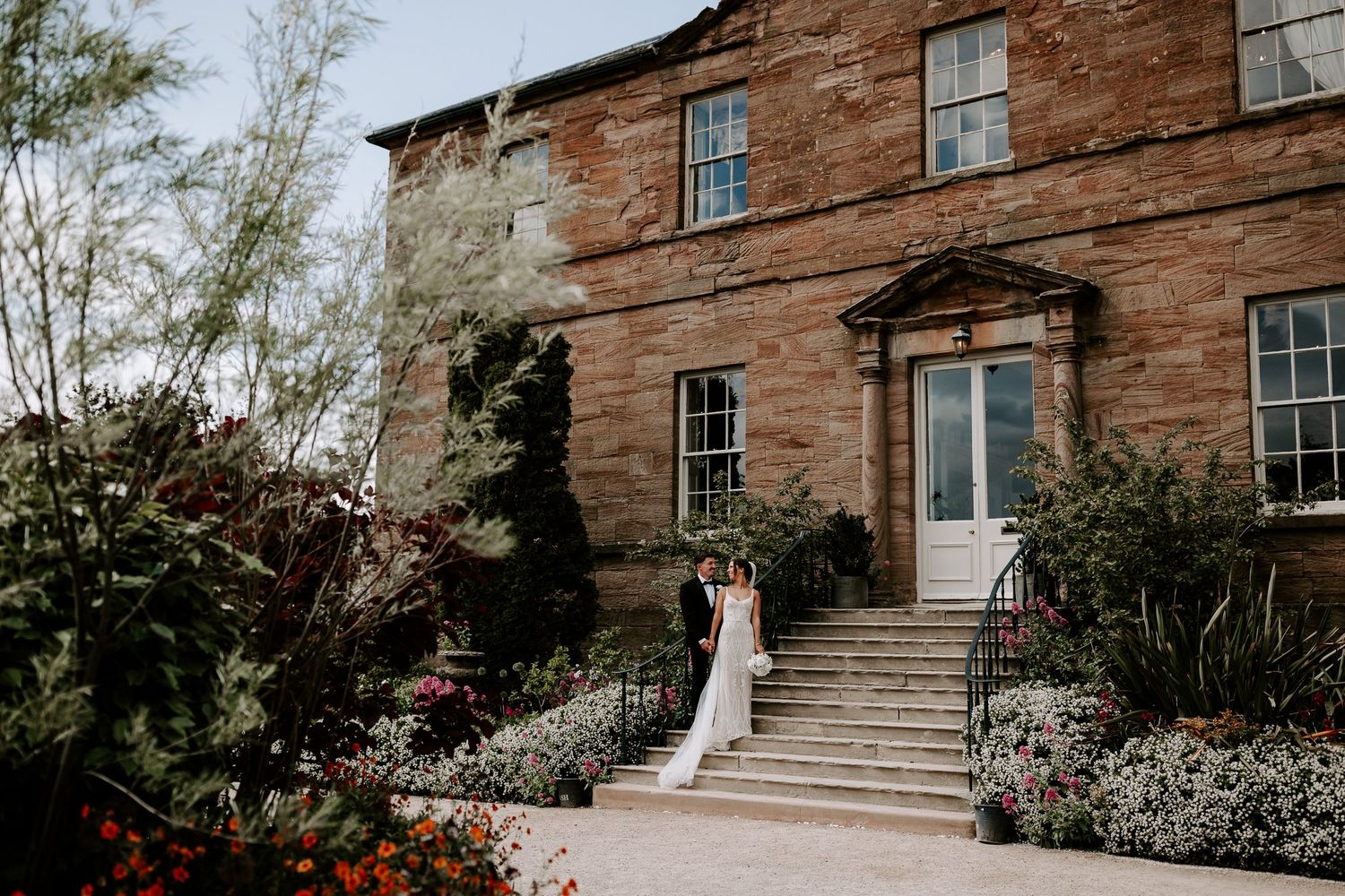 Bride in white gown stands on steps of elegant red brick manor house surrounded by lush gardens.