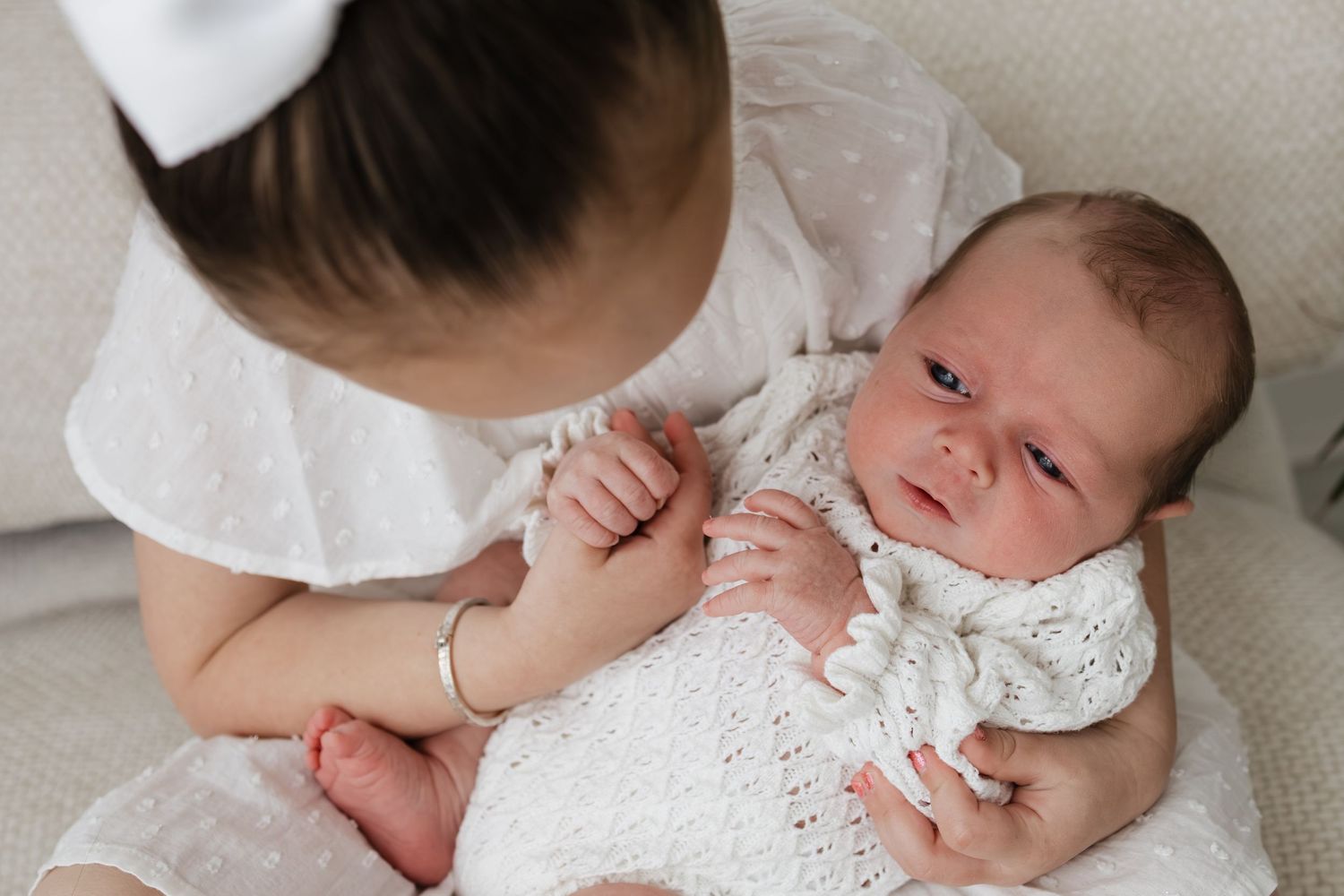 Young girl in white dress gently holds newborn baby wrapped in knit blanket during a soft, tender sibling portrait.