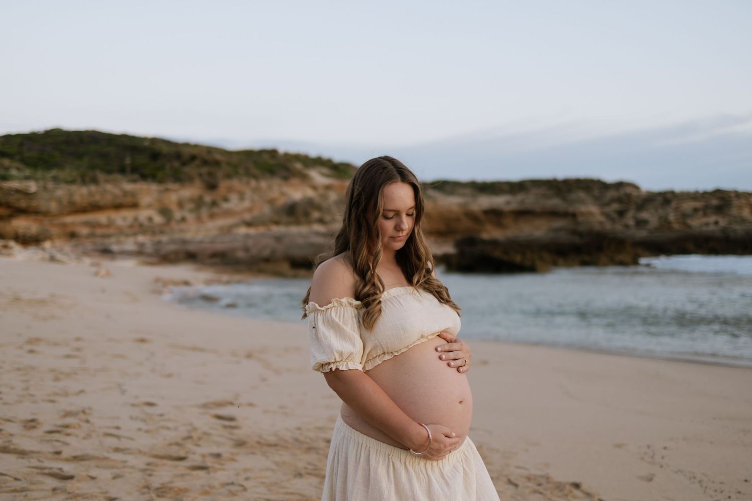 Pregnant woman in white dress cradling baby bump on sandy beach with rocky cliffs and ocean backdrop.