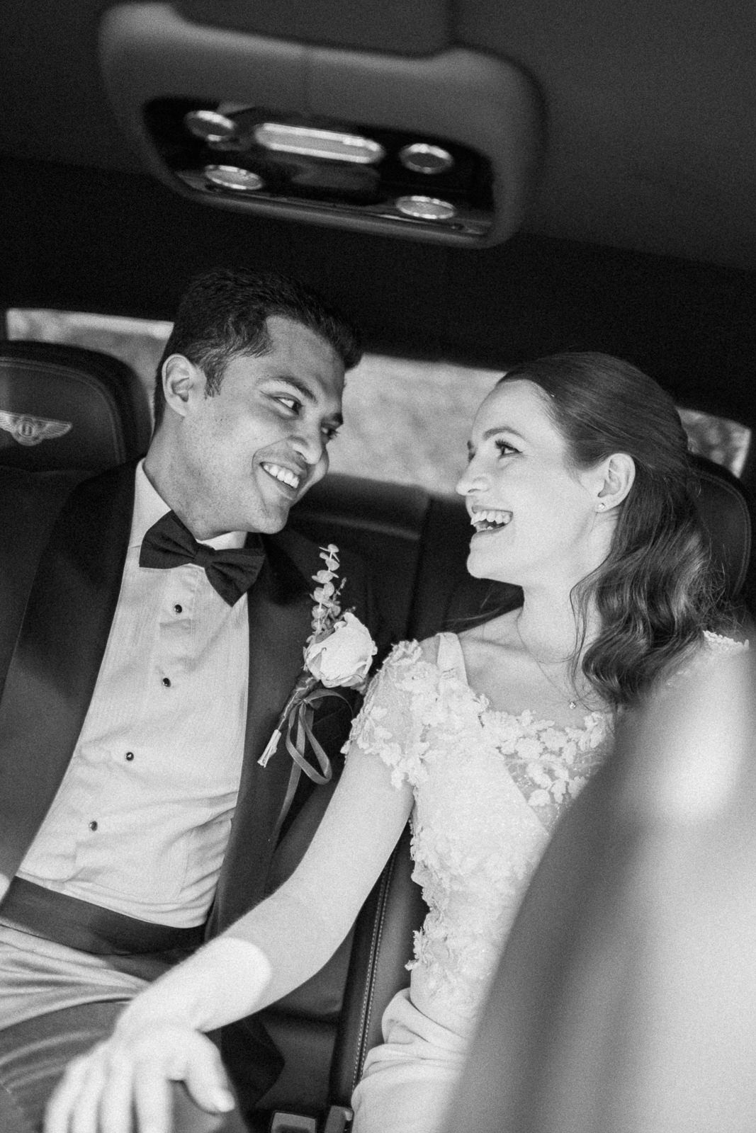 Black and white photo of a smiling bride and groom sharing a joyful moment together in the back of a luxury car.