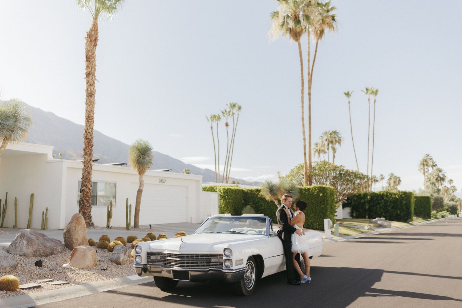 Couple poses by vintage white Cadillac convertible in Palm Springs with palm trees and mountains backdrop.