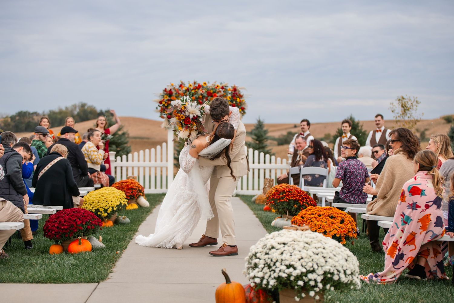 Couple shares first kiss at outdoor fall wedding ceremony with pumpkins, mums, and floral arch under cloudy sky.