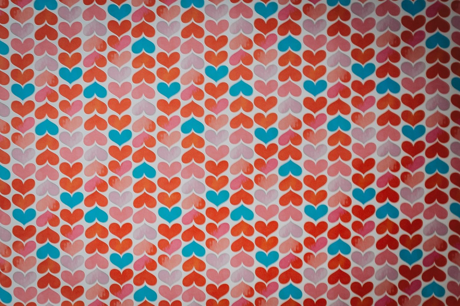 Geometric pattern of red, pink and turquoise heart shapes arranged in vertical stripes on a light background.