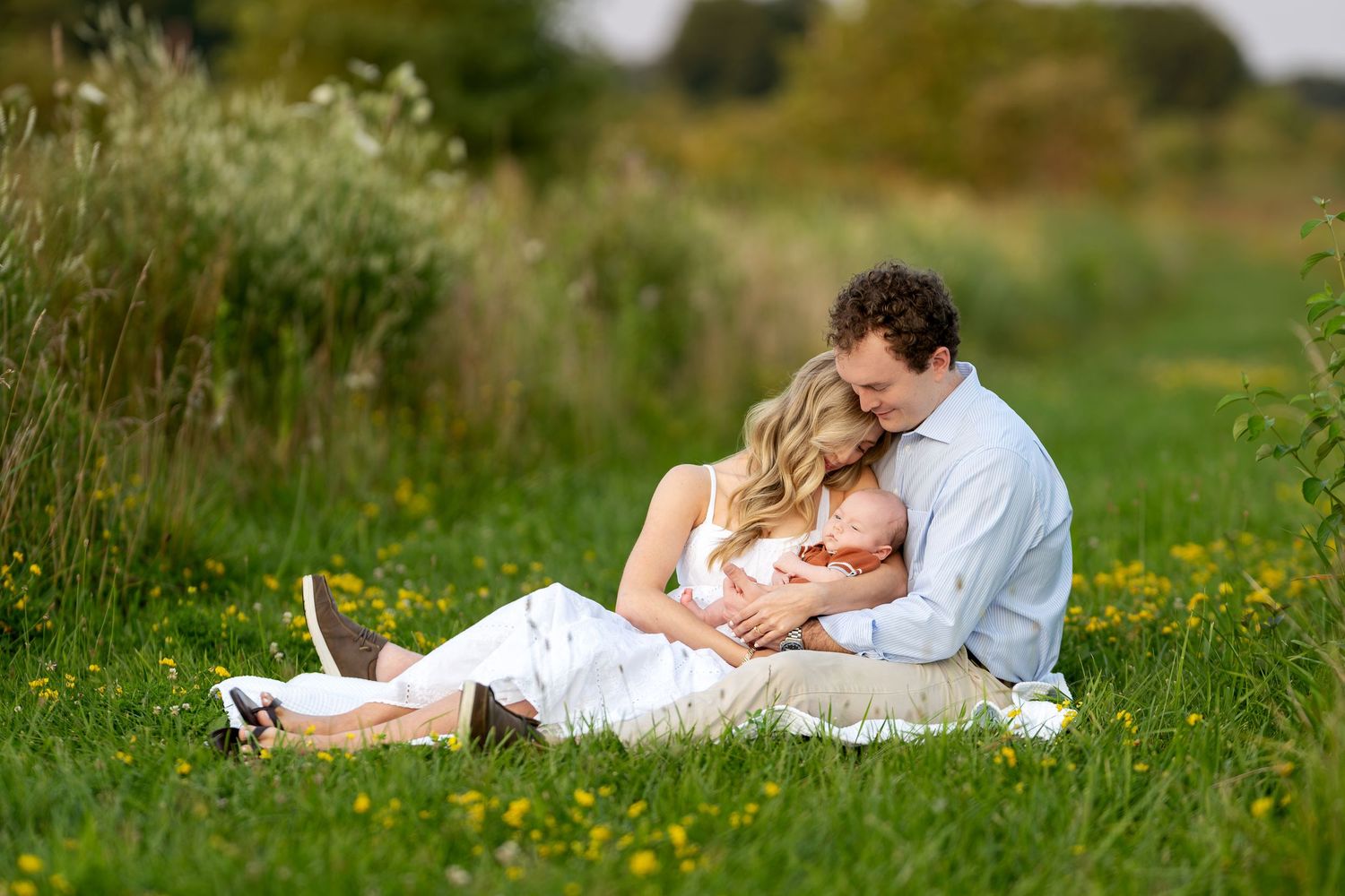 A couple sits in a lush green meadow with wildflowers, tenderly cradling their newborn baby together.