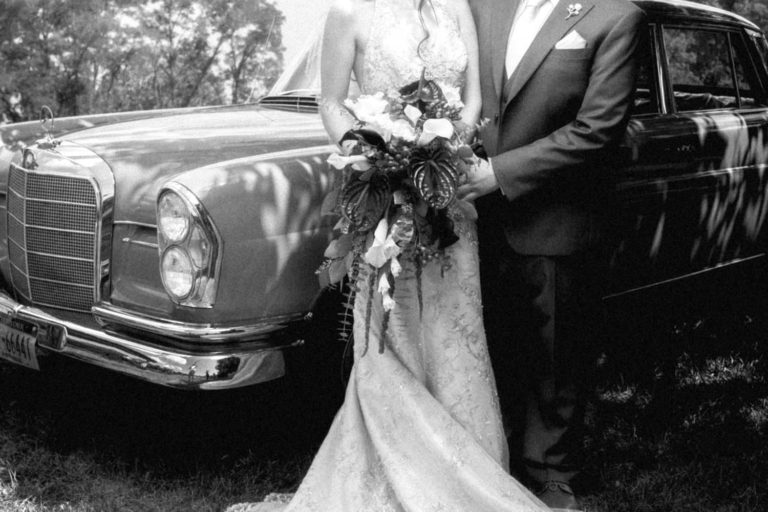 Black and white photo of a bride and groom standing beside a vintage Mercedes-Benz car, bride holding a floral bouquet.