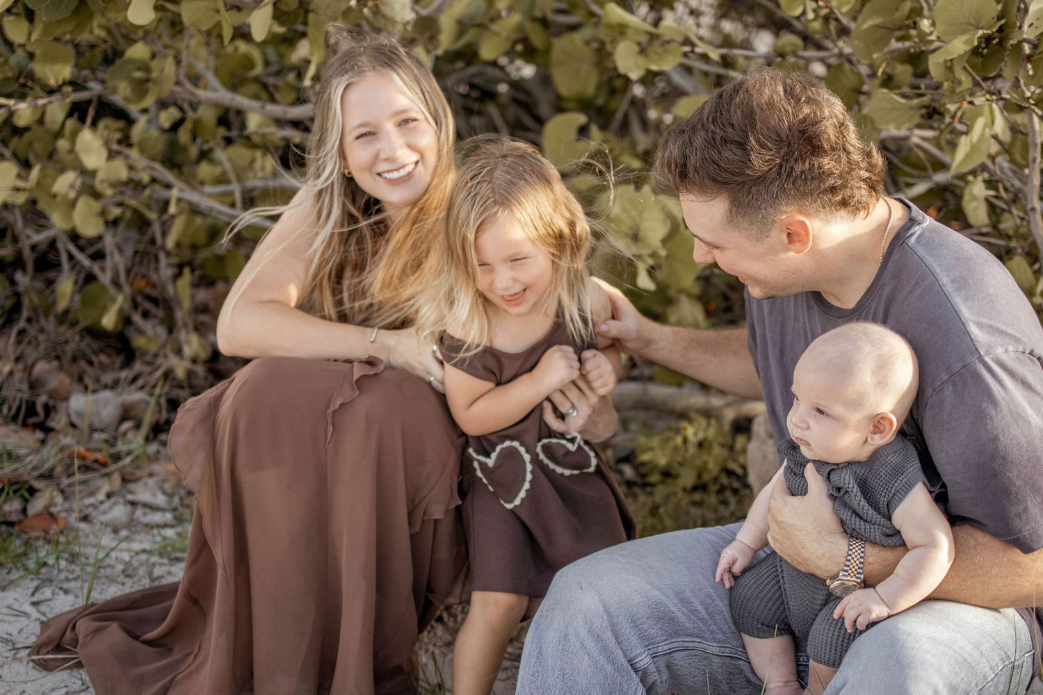 Happy family with two children smiling outdoors near lush green plants on a sandy beach.