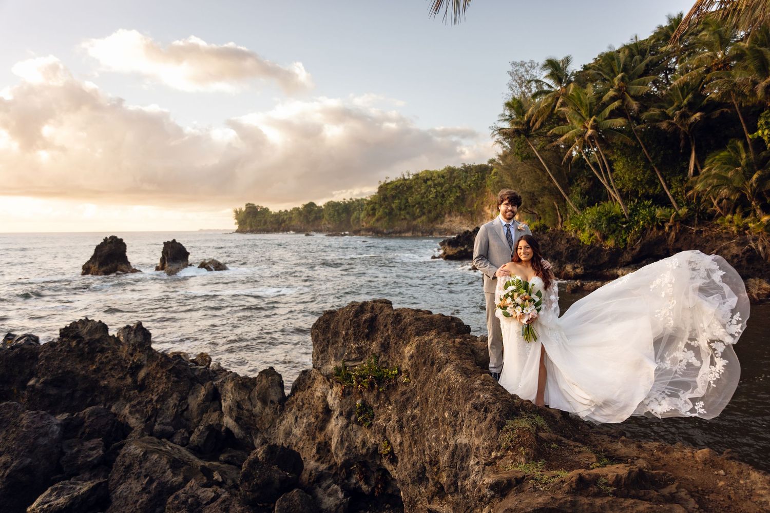 Bride and groom pose on lava rocks at sunset with tropical coastline and palm trees.