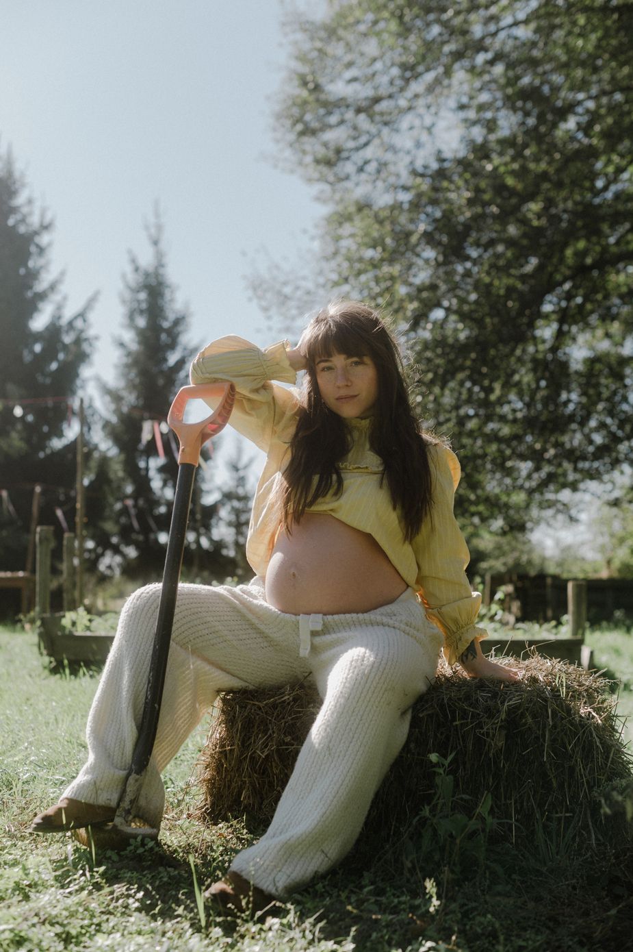 Pregnant woman in yellow top sits on hay bale outdoors, holding a garden tool on a sunny day.