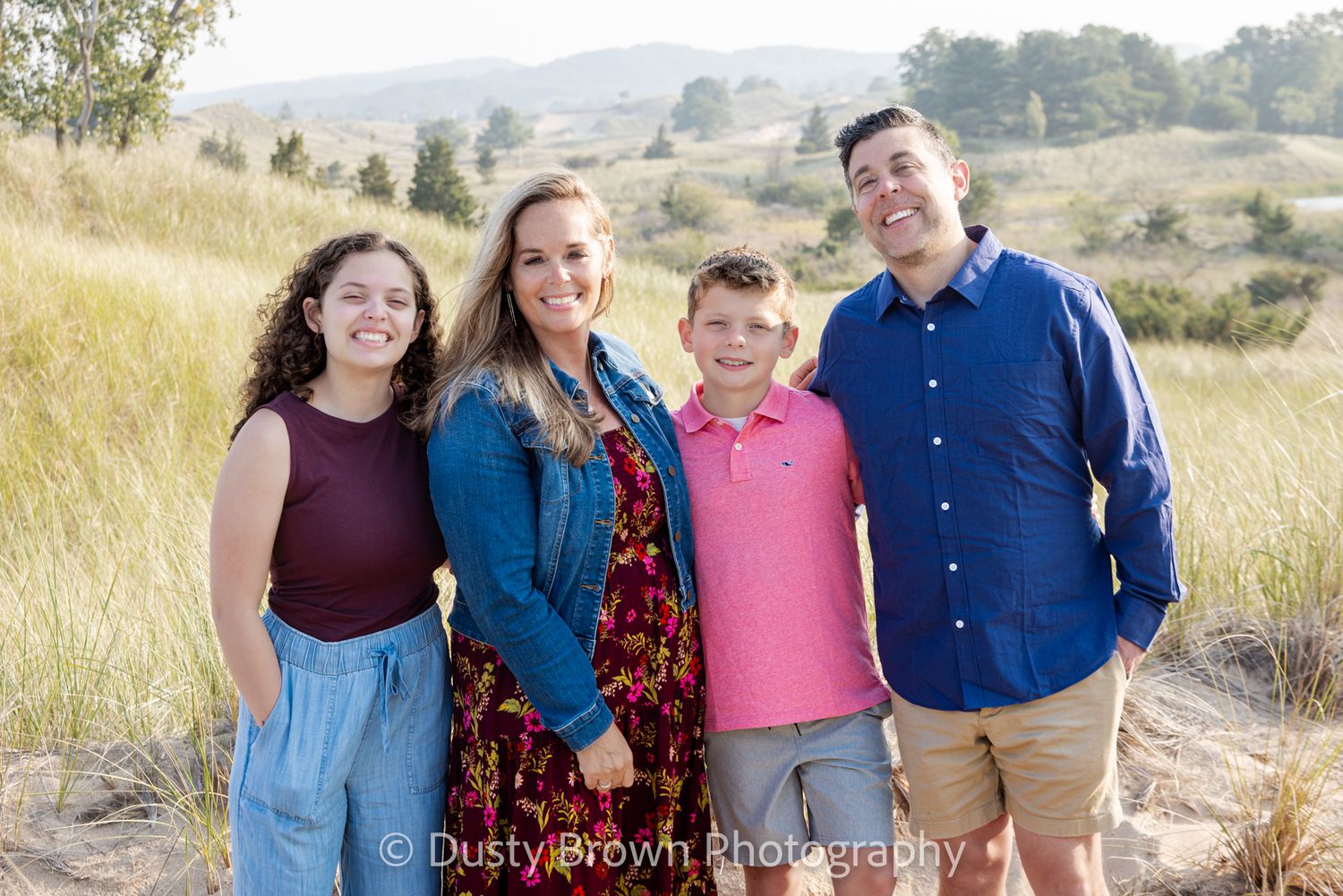 A group of four stands together smiling in tall grass with rolling hills and mountains in the background.